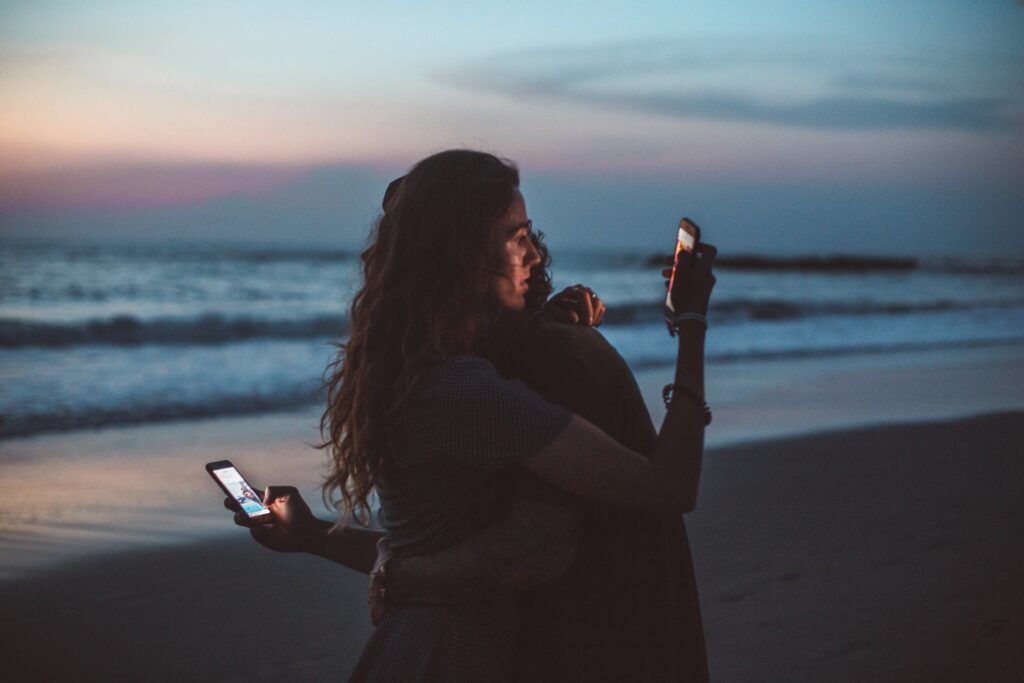 couple hugging and using smartphone near sea on sunset