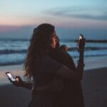 couple hugging and using smartphone near sea on sunset